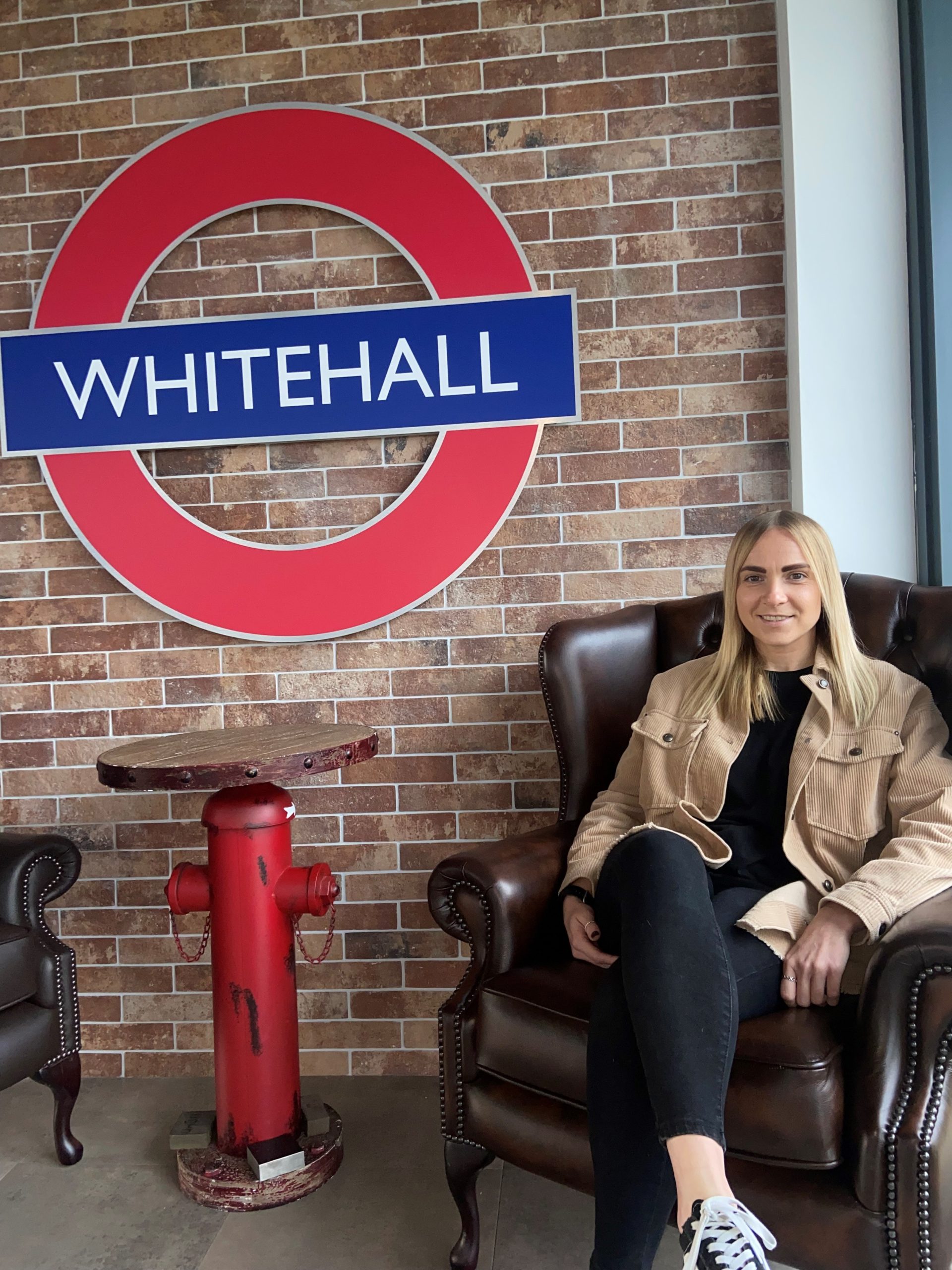 A lady sitting in a chair with thew Whitehall logo on a wall behind her